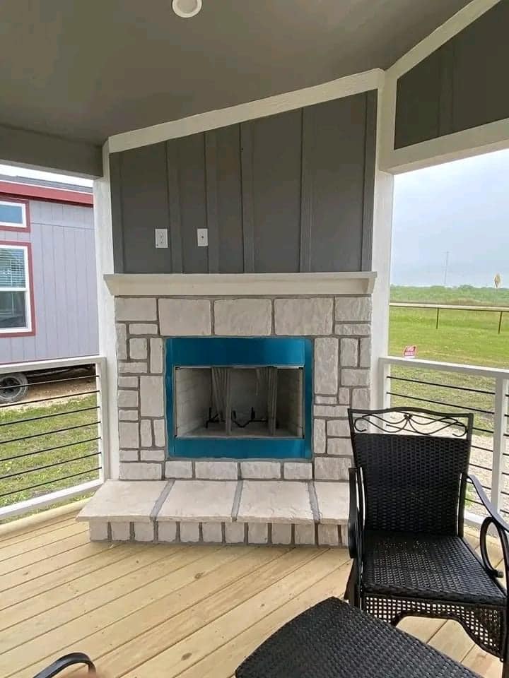 Covered patio with a stone fireplace featuring a bright blue accent, surrounded by black wicker chairs on a wooden deck, overlooking grassy fields.