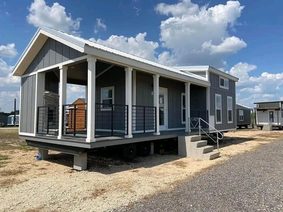 A modern tiny house with grey siding and white trim sits raised on blocks under a bright, blue sky with scattered clouds. A small porch and steps add charm.