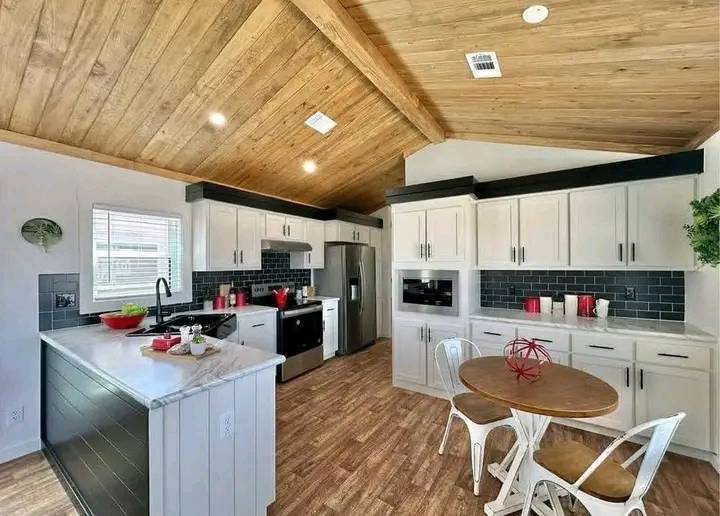 Modern kitchen with vaulted wooden ceiling, white cabinets, stainless steel appliances, and a round wooden table with two chairs. Cozy and inviting.