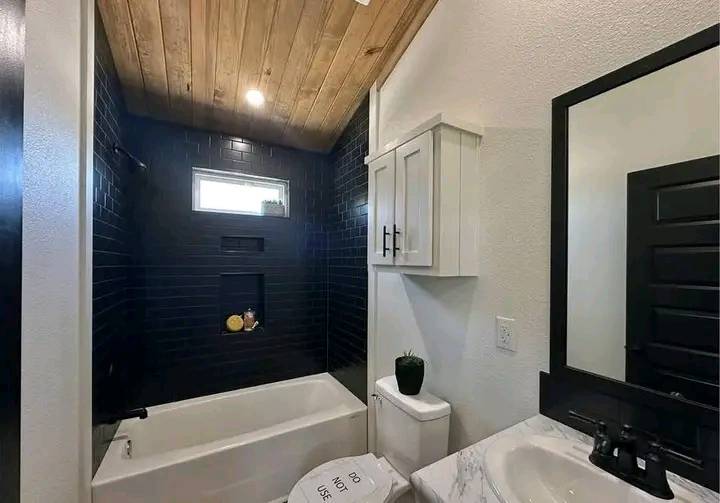 A modern bathroom features a wooden ceiling, dark tiled walls, and a white bathtub. A small window provides light, creating a sleek, cozy atmosphere.