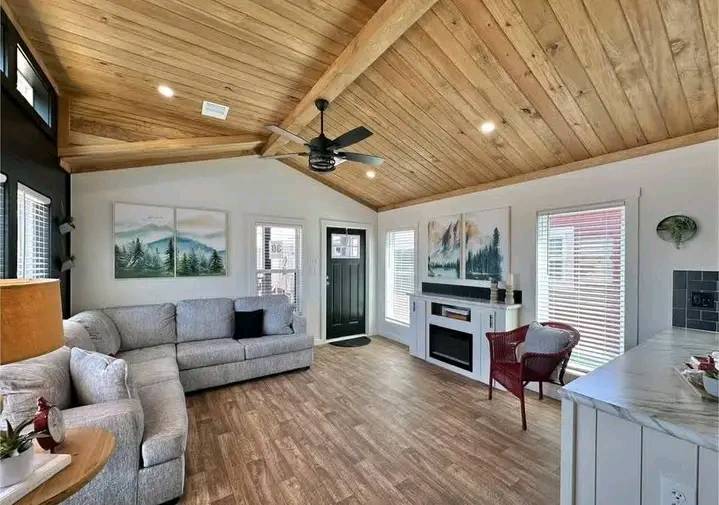 Cozy living room with wood-paneled ceiling and floor, gray sectional sofa, fireplace, artwork, ceiling fan, and large windows. Bright and inviting.