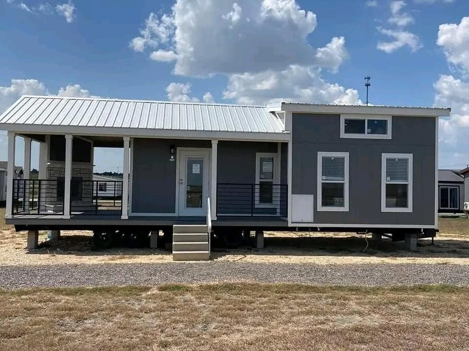 Modern tiny home on stilts with a gray exterior and white roof. It features a small porch, two front windows, steps leading up, and a clear blue sky above.