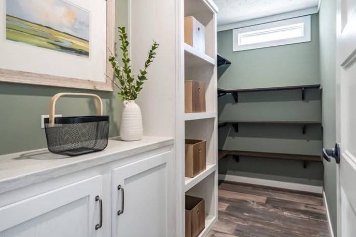 A modern pantry with green walls, wooden shelving, and a countertop with a black basket and vase. Natural light enters through a high window, creating an airy feel.