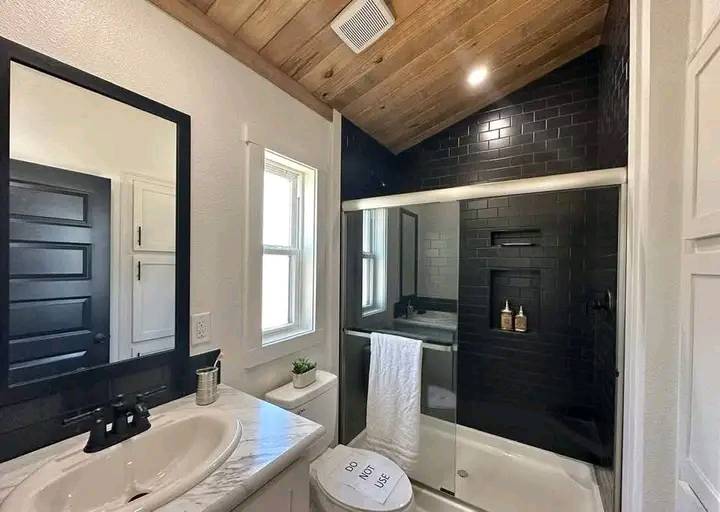 Modern bathroom with wood-paneled ceiling and skylight. Features black-tiled shower, white sink, mirror, window, and wood accents. Calm, minimalist tone.