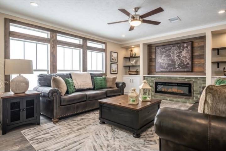 Cozy living room with a brown leather sofa, green pillows, large windows, and a ceiling fan. A fireplace is surrounded by shelves and wall art.