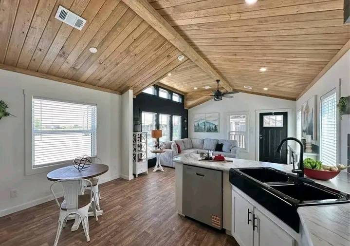 Spacious living area with vaulted wooden ceiling, cozy gray couch, round dining table, and kitchen island. Sunlight streams through ample windows.