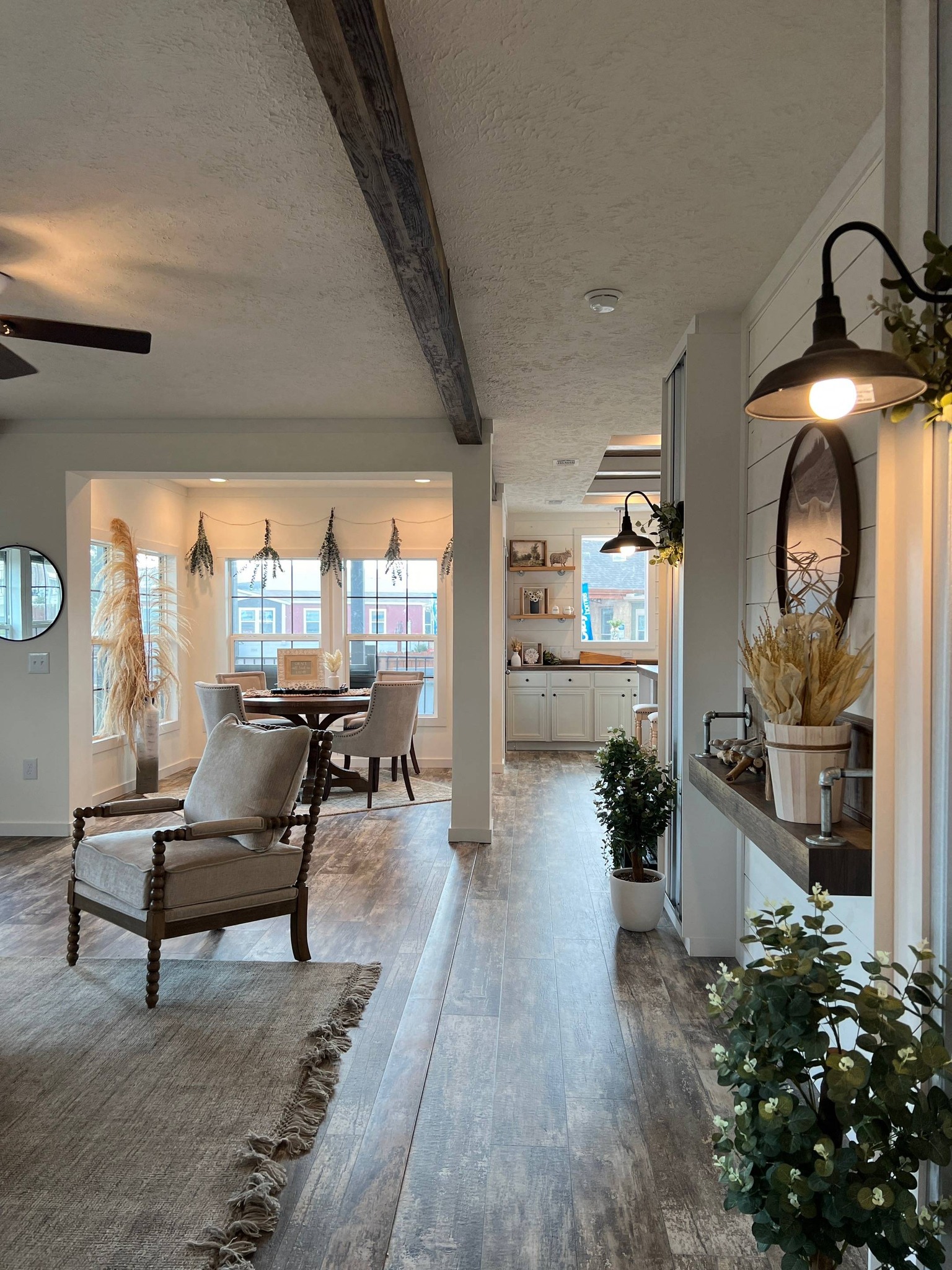Bright, rustic living space with a wooden floor, exposed beam, and cozy decor. A dining area with a round table is near large windows, creating an inviting atmosphere.