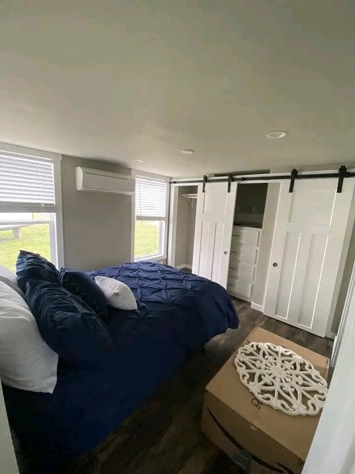 Small bedroom with dark wood flooring, a bed with navy bedding, white pillows, and a decorative box. Sliding barn doors reveal an open closet. Natural light streams through two tall windows, creating a cozy and serene atmosphere.