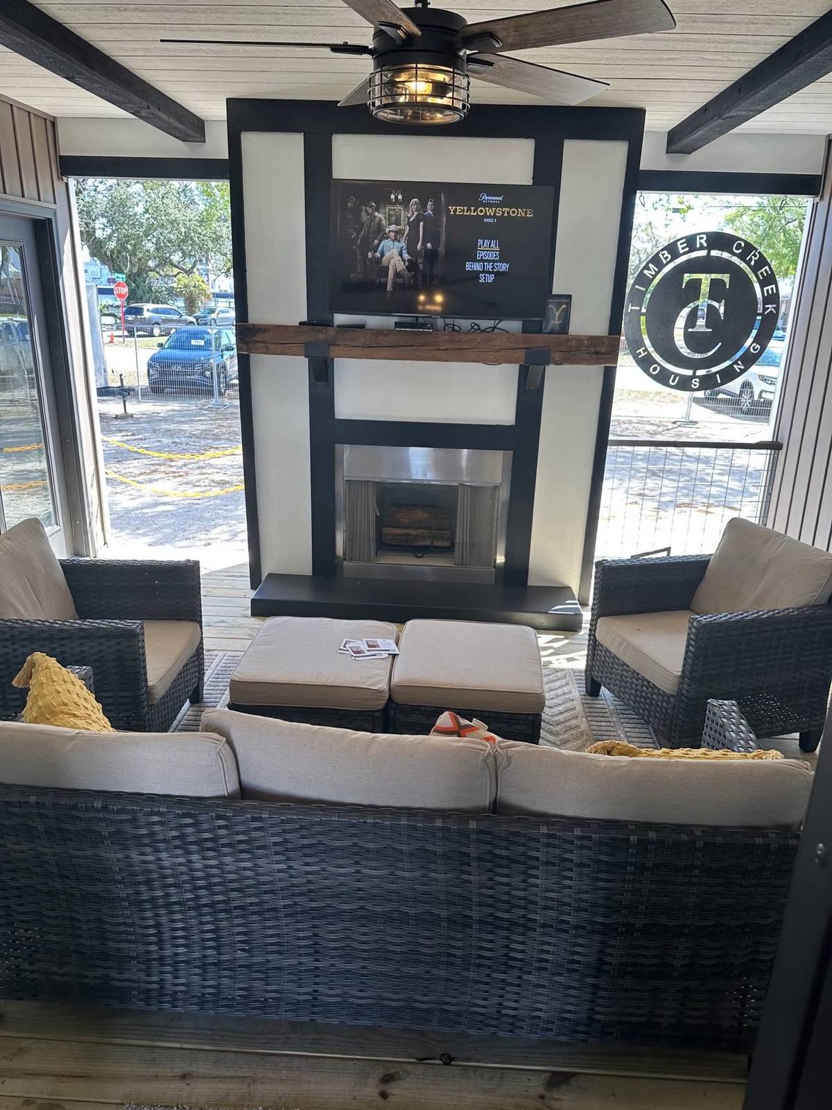 Outdoor seating area with wicker furniture facing a TV showing "Yellowstone." Modern fireplace beneath, ceiling fan above. Relaxing ambiance.