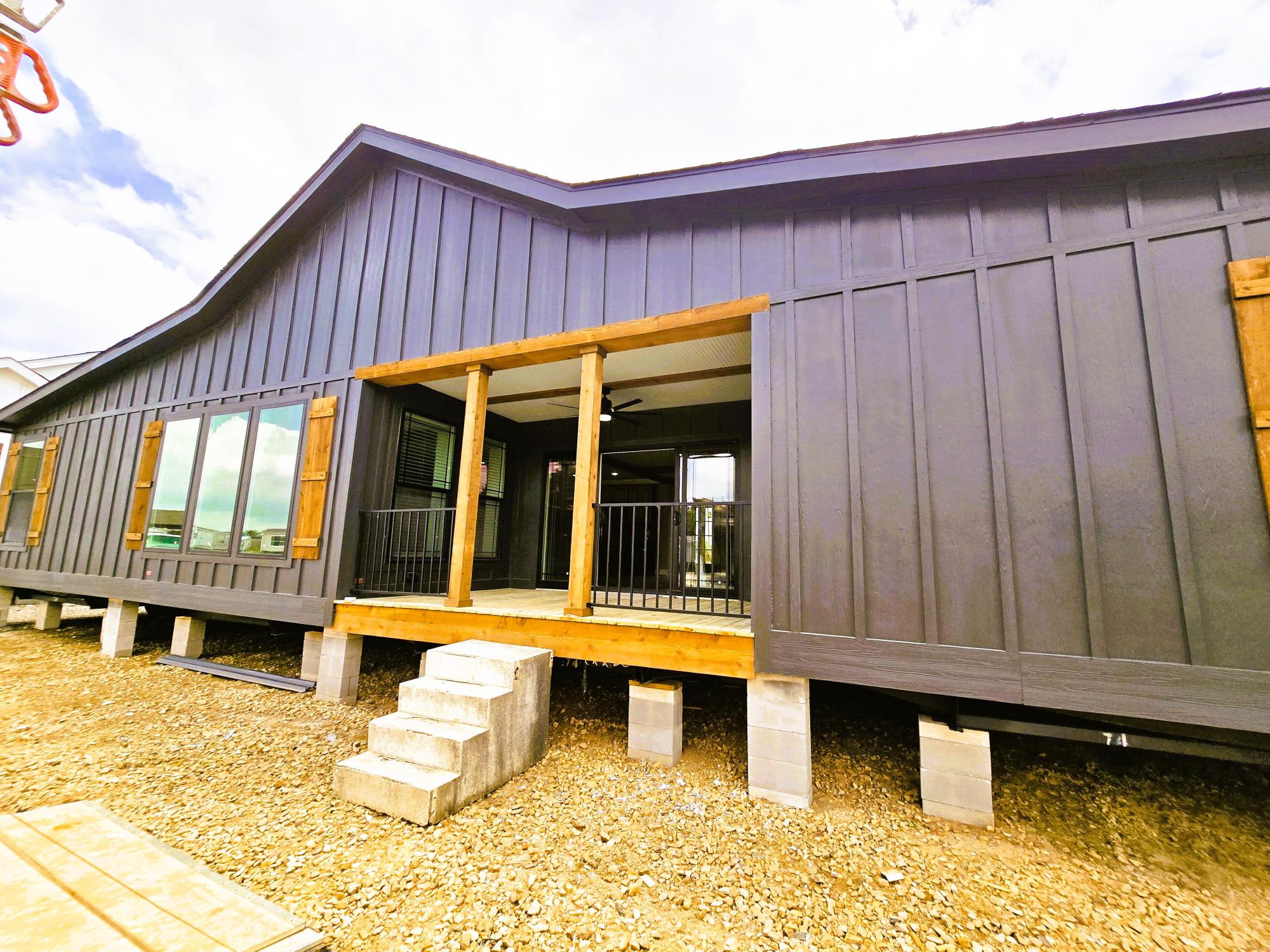 Modern modular home exterior with dark wood paneling and large windows. The small porch has wooden beams, cinder block steps, and a minimalist style. Sky above is bright and partly cloudy.