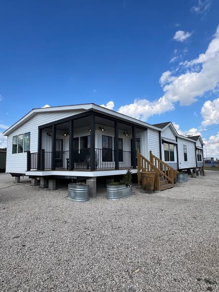A modern, white mobile home with a black-trimmed porch sits elevated on gravel under a blue sky with clouds. The scene is bright and inviting.