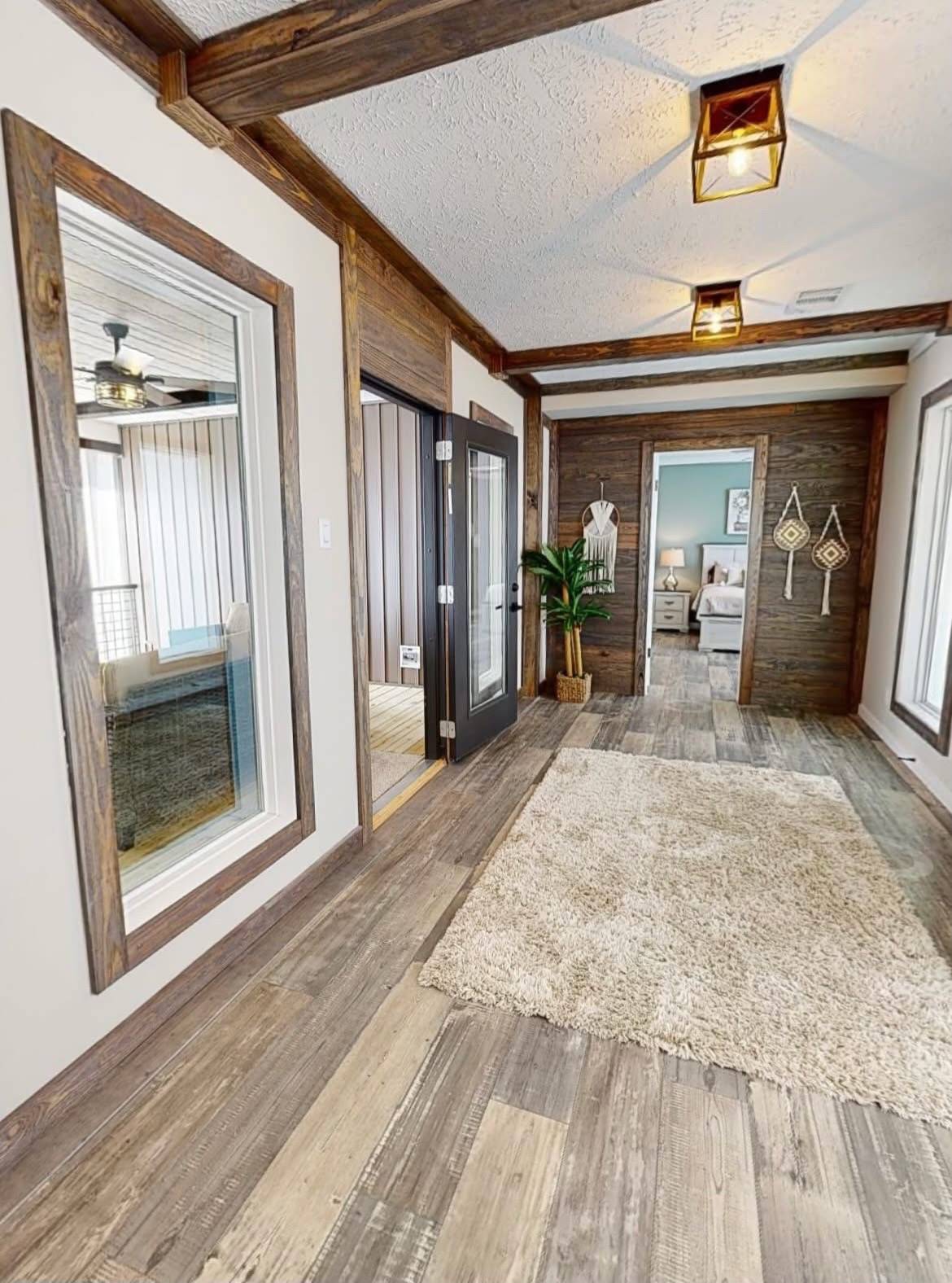 Hallway with rustic wood flooring and exposed beams, featuring a beige rug. A door leads to a bedroom with a coastal decor theme. Warm and inviting atmosphere.