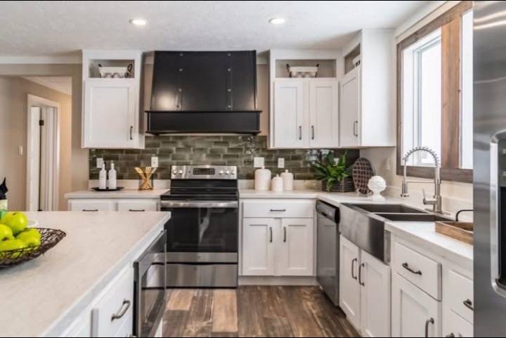 Modern kitchen with white cabinets, black appliances, and a farmhouse sink. Green tile backsplash and wood floors add warmth. Apples in a bowl suggest freshness.