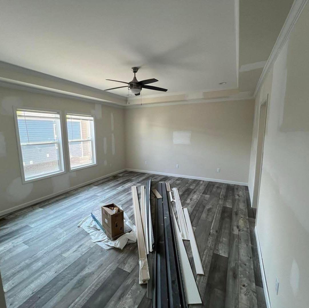 Spacious, empty room with new wood flooring, beige walls, two windows, a ceiling fan, and construction materials, including planks and a box, on the floor.