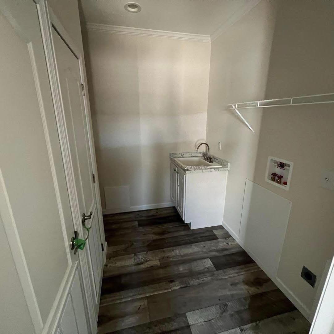 A small laundry room with dark wood flooring, white walls, and a white cabinet with a sink. A shelf is mounted on the right wall, evoking a minimalist feel.