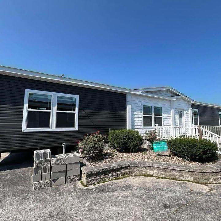 A modern manufactured home with dark siding and white trim, bordered by a small garden and ramp, sits under a clear blue sky. A stack of cinder blocks is visible nearby.
