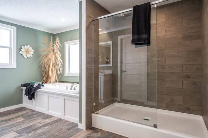 Spacious bathroom with a modern glass shower, a bathtub adorned with pampas grass, and a green wall. Natural light streams through two windows.