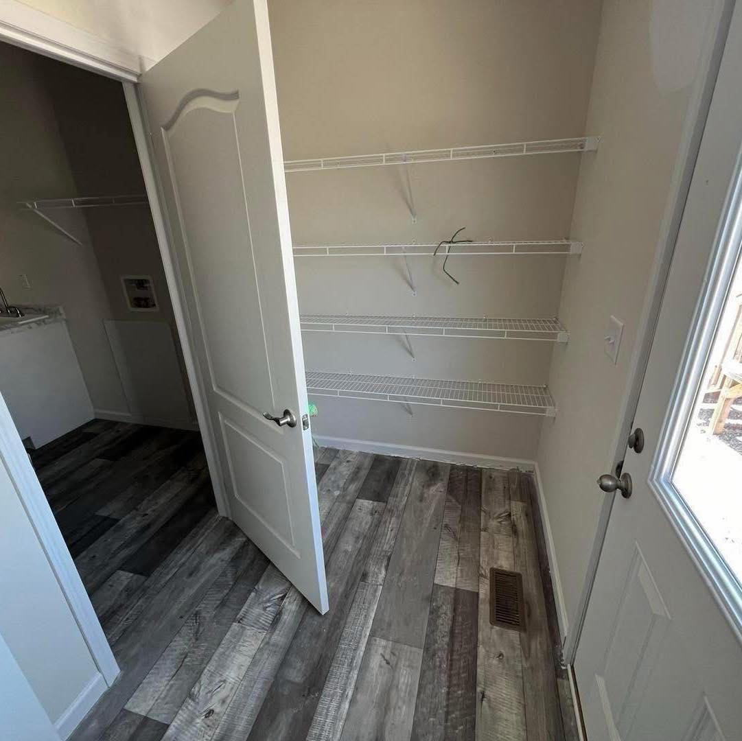 Room with open door and wooden floor, featuring three empty white wire shelves on a beige wall. Air vent on the floor; bright and clean ambiance.