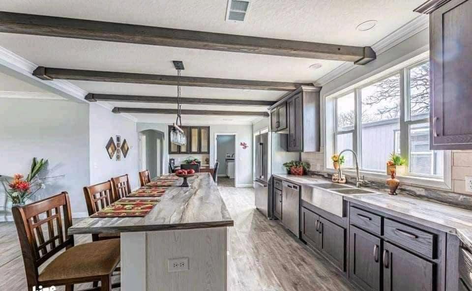 Spacious kitchen with wooden beams, light granite countertops, dark cabinetry, and large windows. A long island with chairs and red decor create a warm, inviting tone.