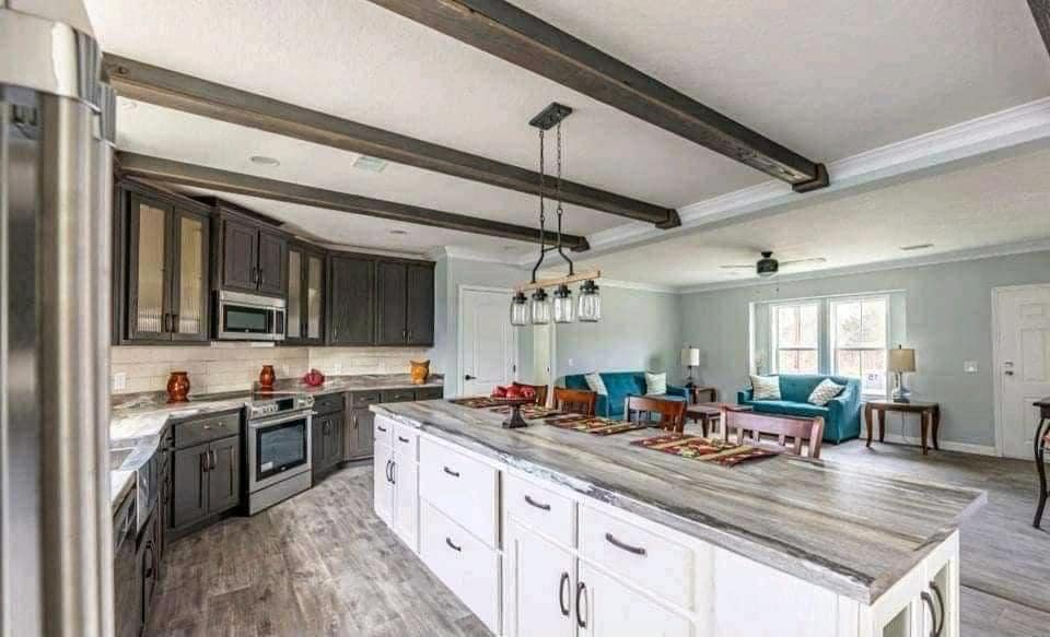 Spacious kitchen with dark wood cabinets, a large island with white drawers, and pendant lights. Open living area with teal sofas and bright windows.