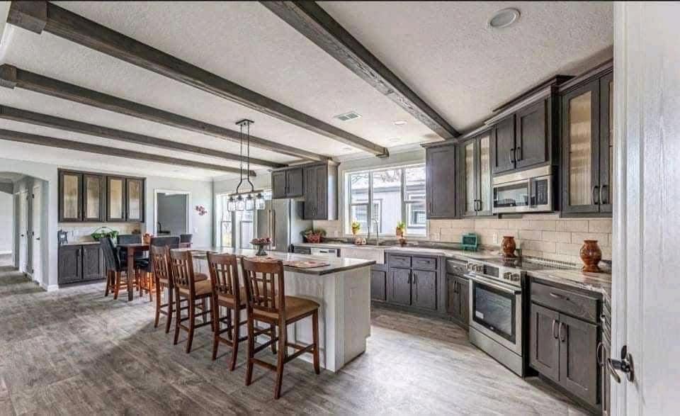 Spacious kitchen with gray cabinets, light wood flooring, and a central island with barstools. Beamed ceiling and stainless steel appliances add modern elegance.
