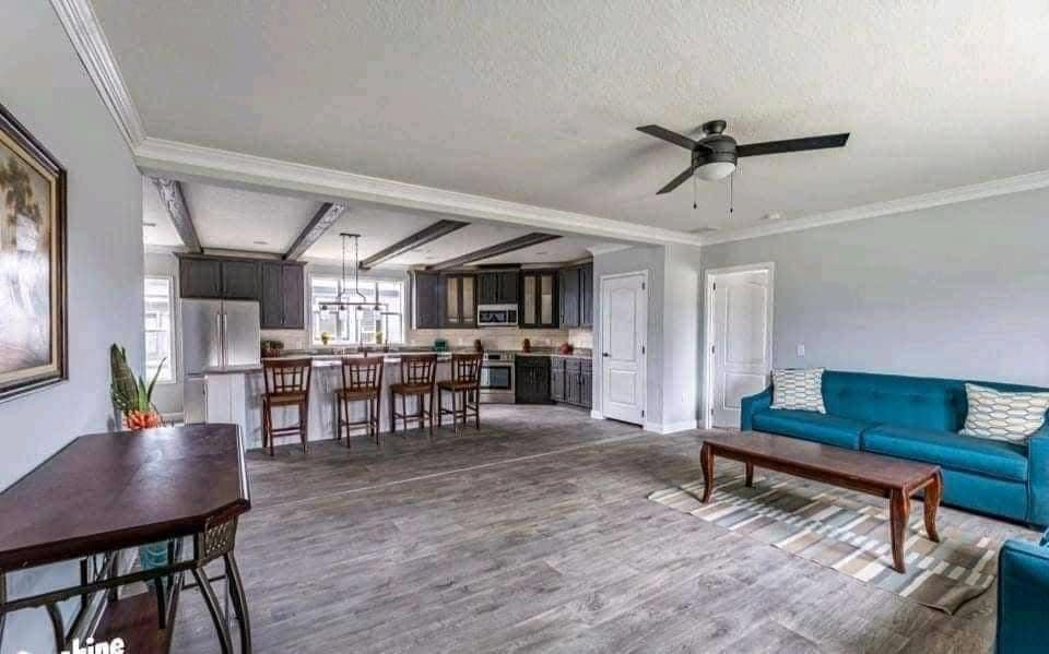 Spacious modern living area with gray wood flooring, teal sofas, and a ceiling fan. An open kitchen with bar stools and dark cabinets is visible in the background.