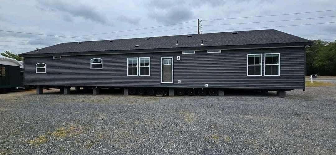 A large, gray, single-story manufactured home on wheels sits on a gravel lot under a cloudy sky. It has multiple windows and a central door.