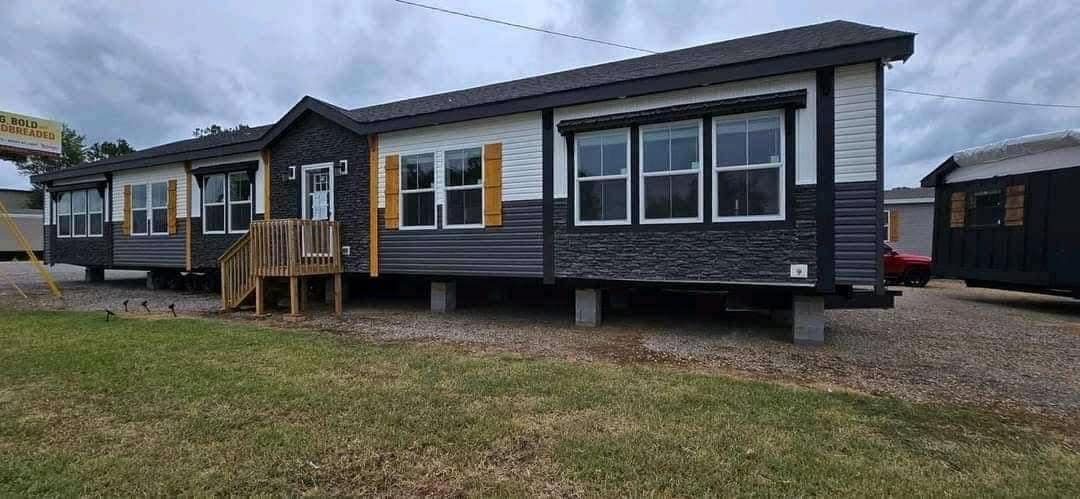 A modern modular home with dark siding, white trim, and large windows sits elevated on blocks. A small wooden porch leads to the front door. Overcast sky.
