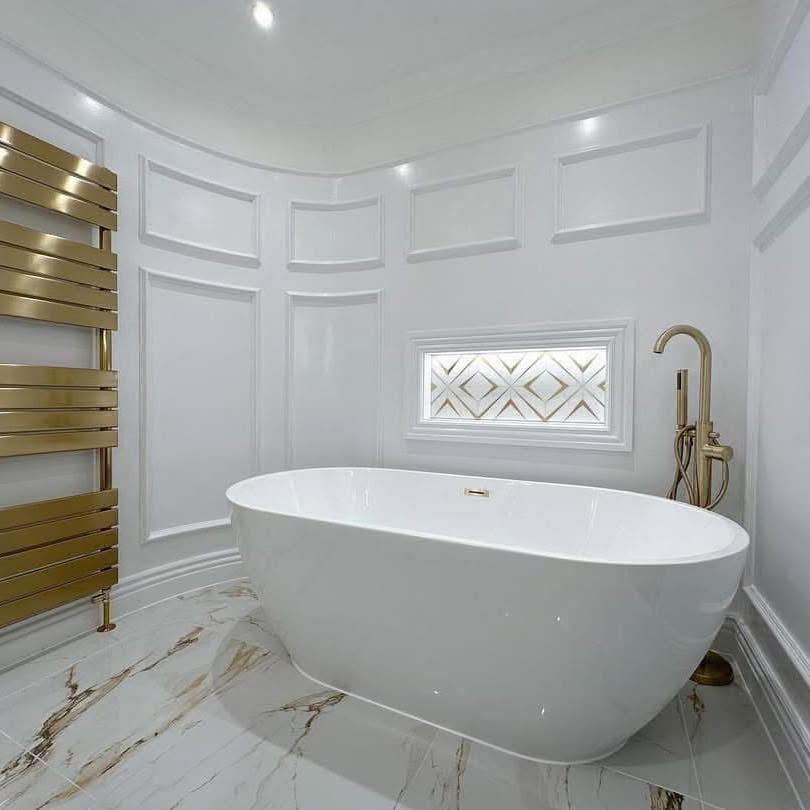 Luxurious bathroom with a sleek white freestanding tub, gold fixtures, marble floor, white paneled walls, and a small decorative window.