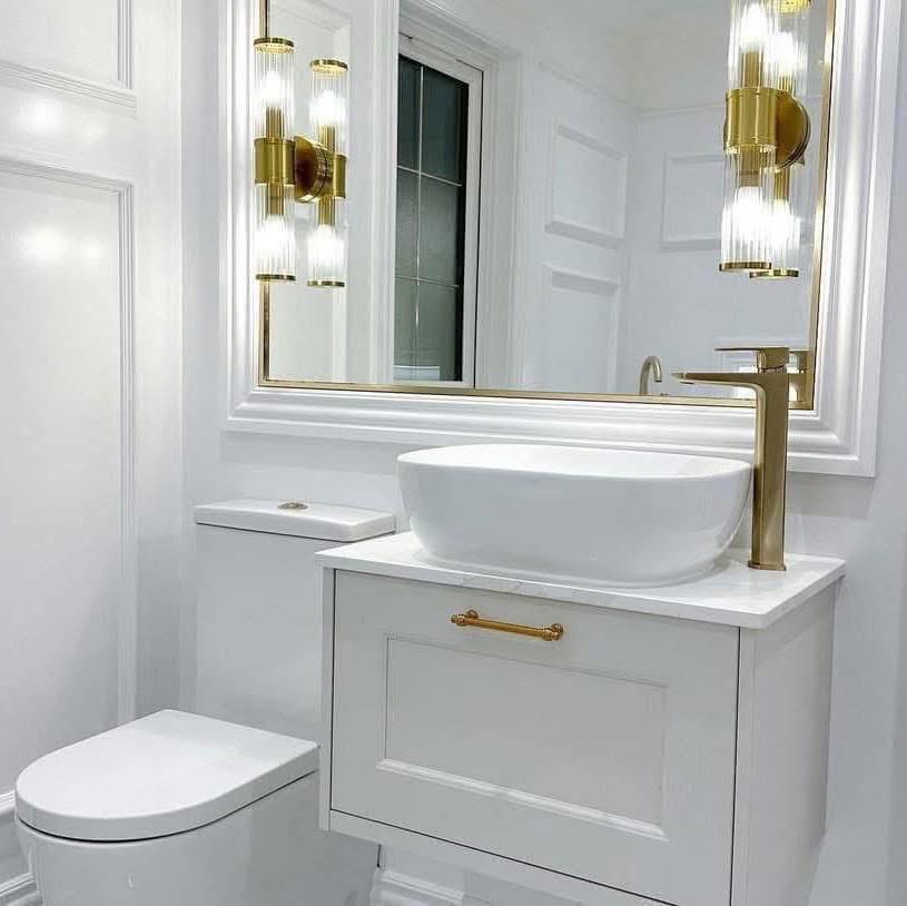 Elegant white bathroom with gold accents featuring a sleek vessel sink, large mirror, modern sconces, and a compact toilet, conveying a luxurious feel.