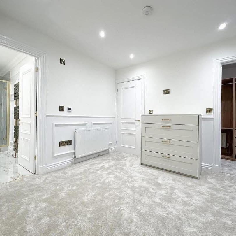 A bright, minimalist room with soft beige carpeting, white walls, and recessed lighting. A white dresser and radiator complement the clean, modern aesthetic.