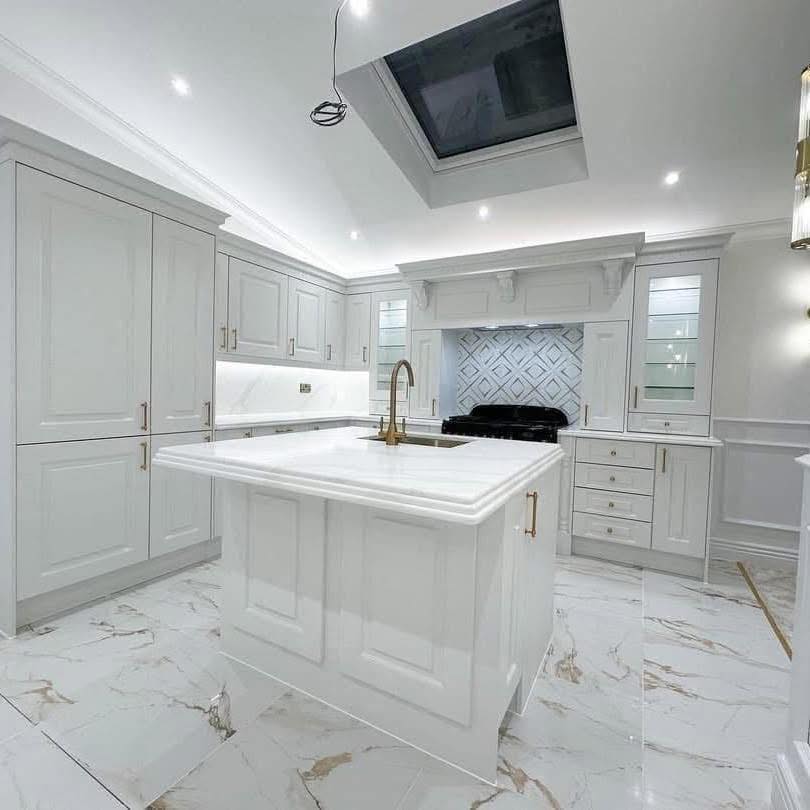 Spacious white kitchen with high ceiling, central island with gold faucet, marble floor, skylight, and patterned backsplash. Bright and modern ambiance.