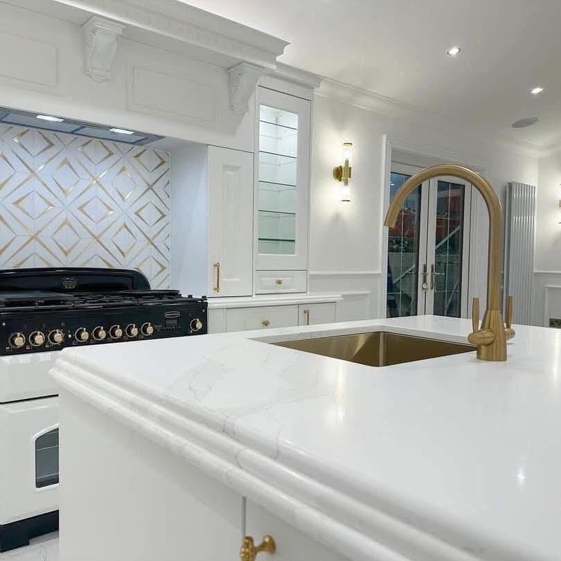 Elegant white kitchen with marble countertops and a gold faucet. A black stove is against a wall with a geometric-patterned backsplash, creating a modern and luxurious feel.