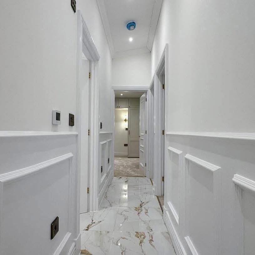 A bright, marble-floored hallway with white paneled walls and recessed lighting. It leads to an open door, conveying a clean and modern aesthetic.
