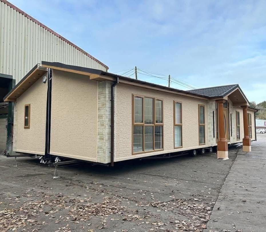 Mobile home with light beige walls, large windows, and a brown roof. It's positioned on a paved area with fallen leaves, next to an industrial building under a clear sky.