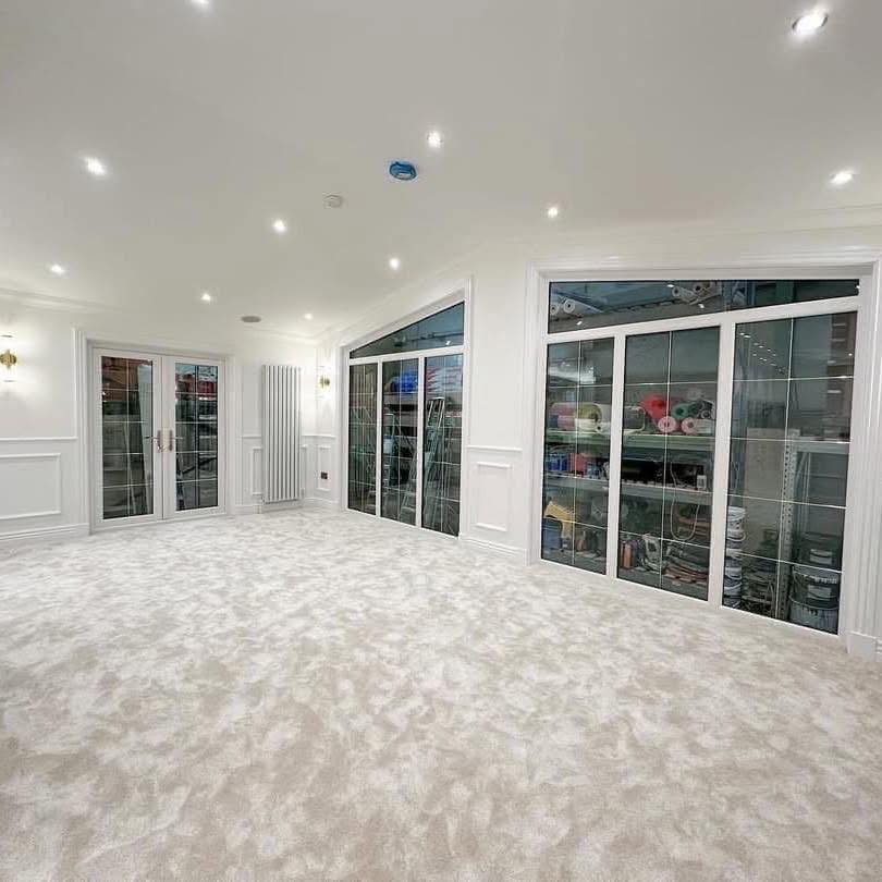 Spacious room with plush white carpet, white walls, and recessed lighting. Large glass cabinets reveal organized storage of colorful fabrics and paint cans.