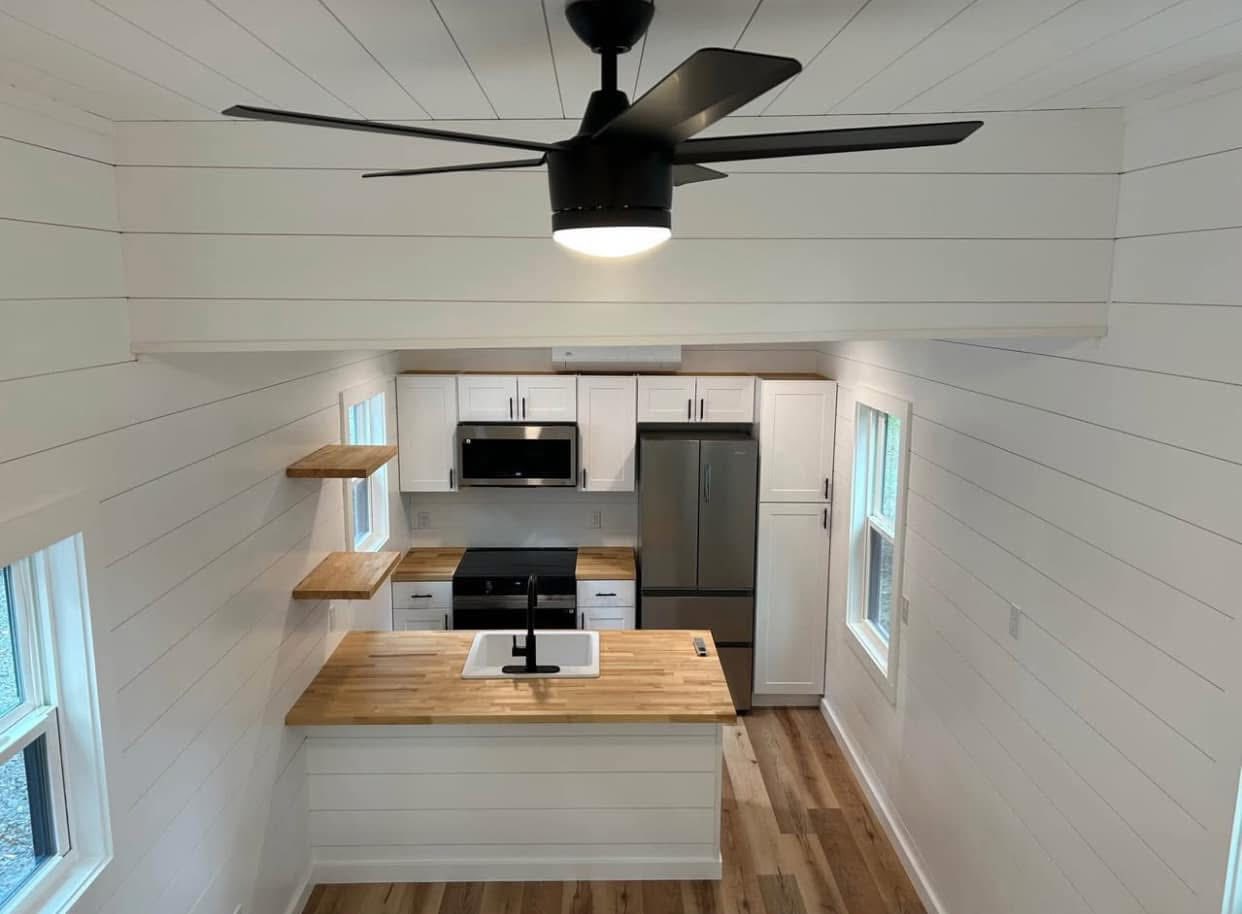 A modern kitchen with white shiplap walls and wood accents. Features a black ceiling fan, wooden countertops, stainless steel appliances, and open shelving.