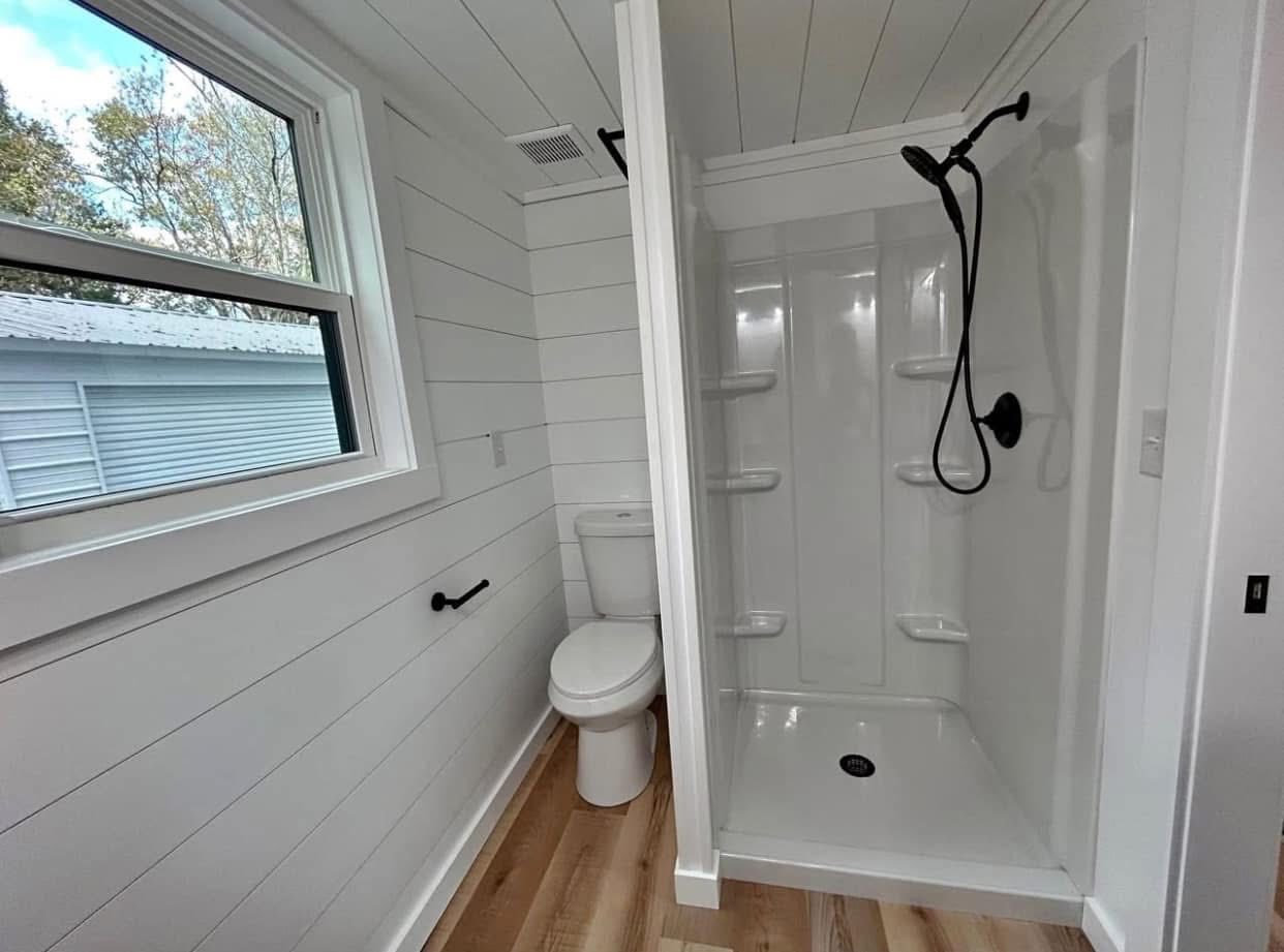 Compact white bathroom with wood flooring, featuring a toilet, open shower with black fixtures, and a window providing natural light. Clean, minimal design.