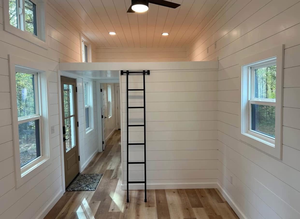 Bright tiny house interior with white shiplap walls, wood flooring, and large windows. A black ladder leads to a loft. Cozy, minimalist design.