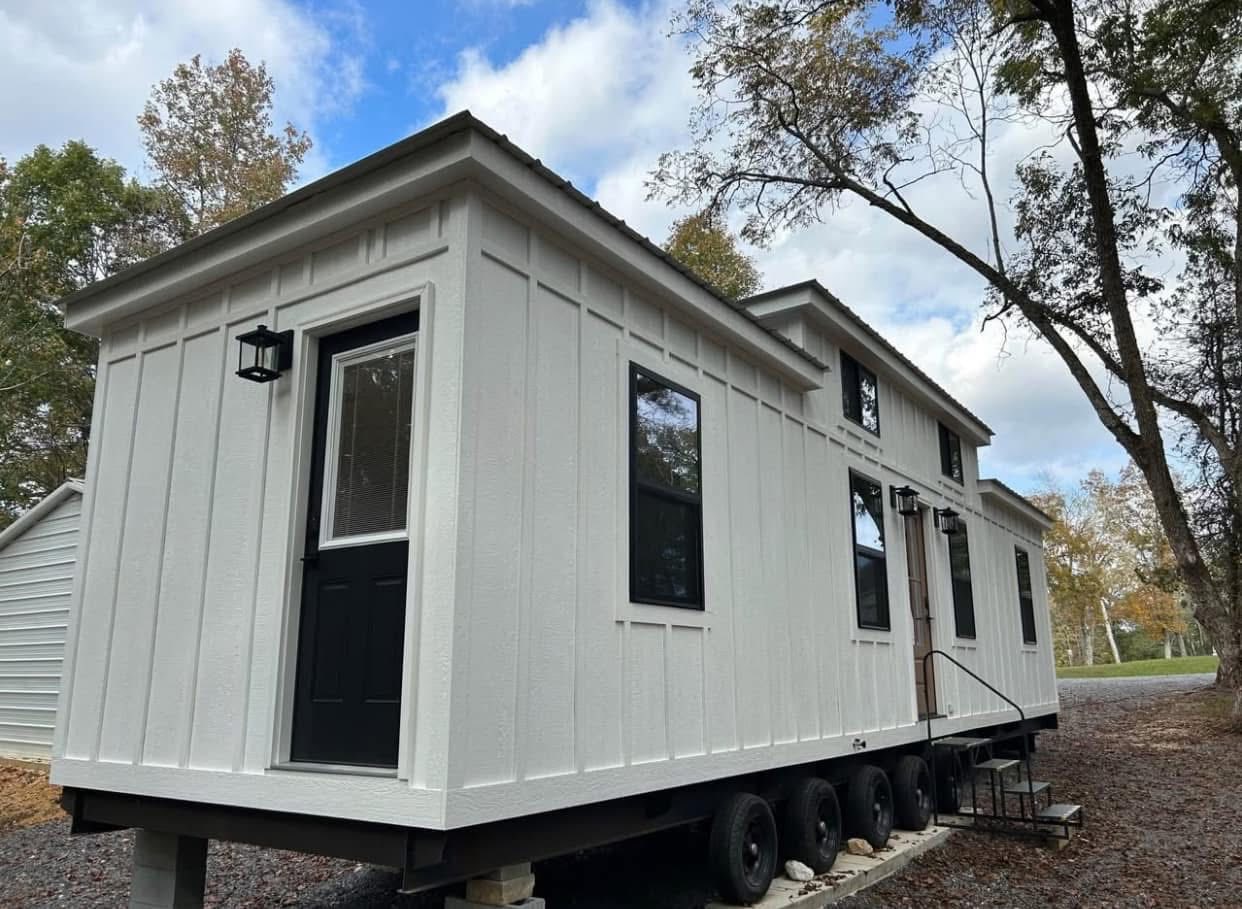 A white tiny house on wheels is parked outdoors under a blue sky with scattered clouds. Surrounded by trees, it conveys a cozy, rustic charm.