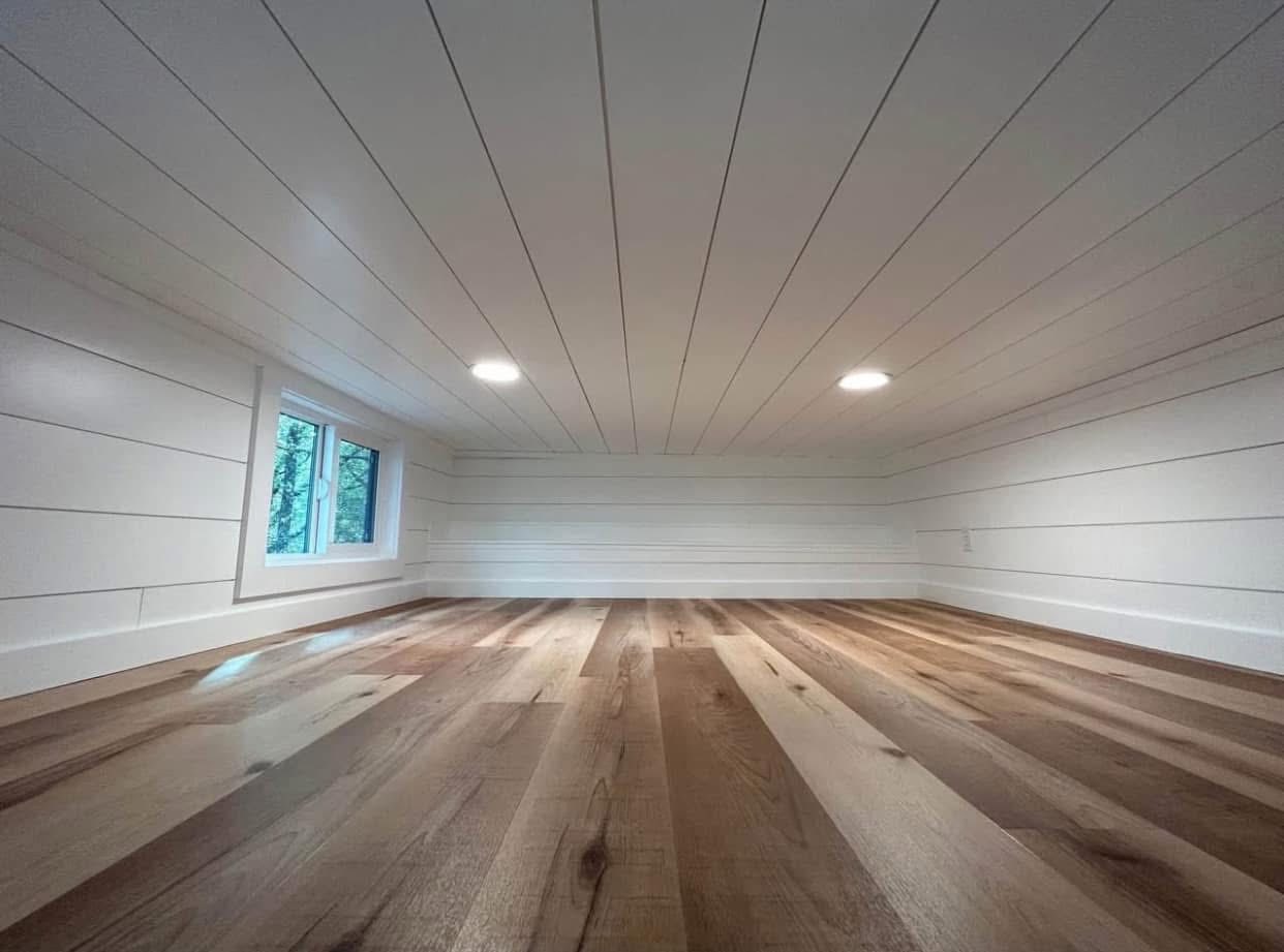 Cozy attic space with white paneled walls and ceiling, featuring a small window showing trees. Warm wooden floors and two circular ceiling lights create a serene vibe.