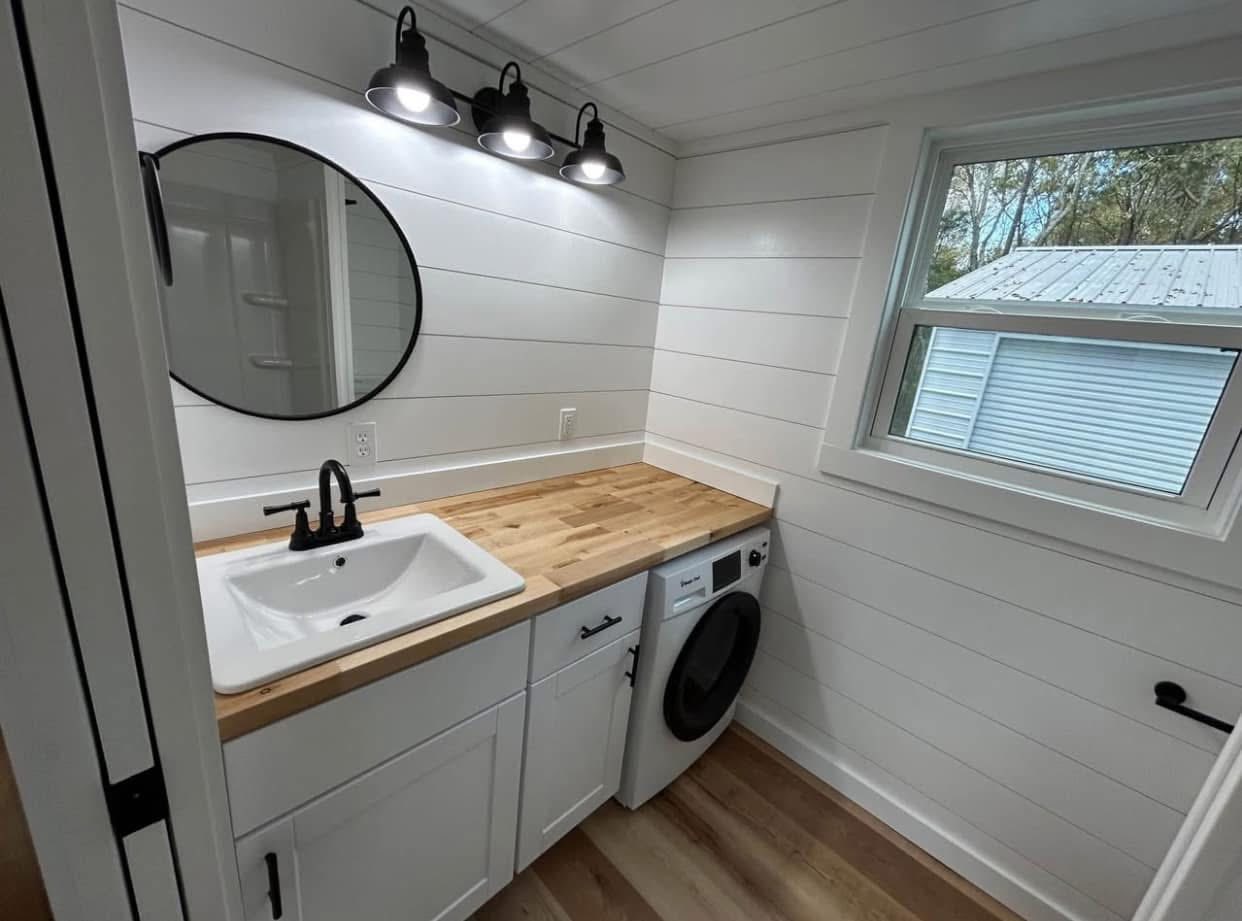 Compact laundry room with white shiplap walls, light wood countertops, a black faucet on a sink, and a round mirror under modern lights; washer below.