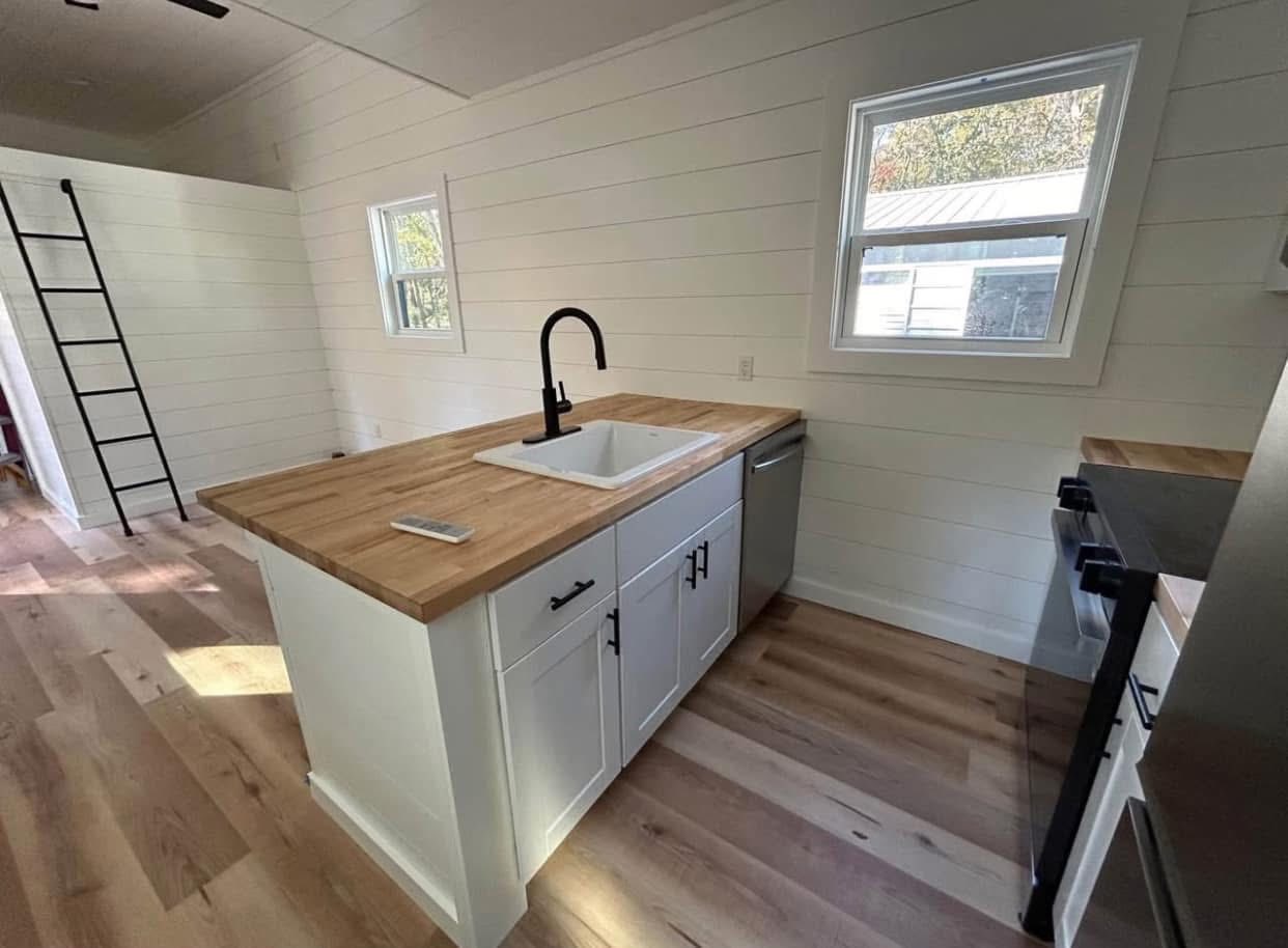 A modern, minimalist kitchen features a wooden countertop island with a white sink and black faucet. Light wood flooring and white shiplap walls create an airy feel.