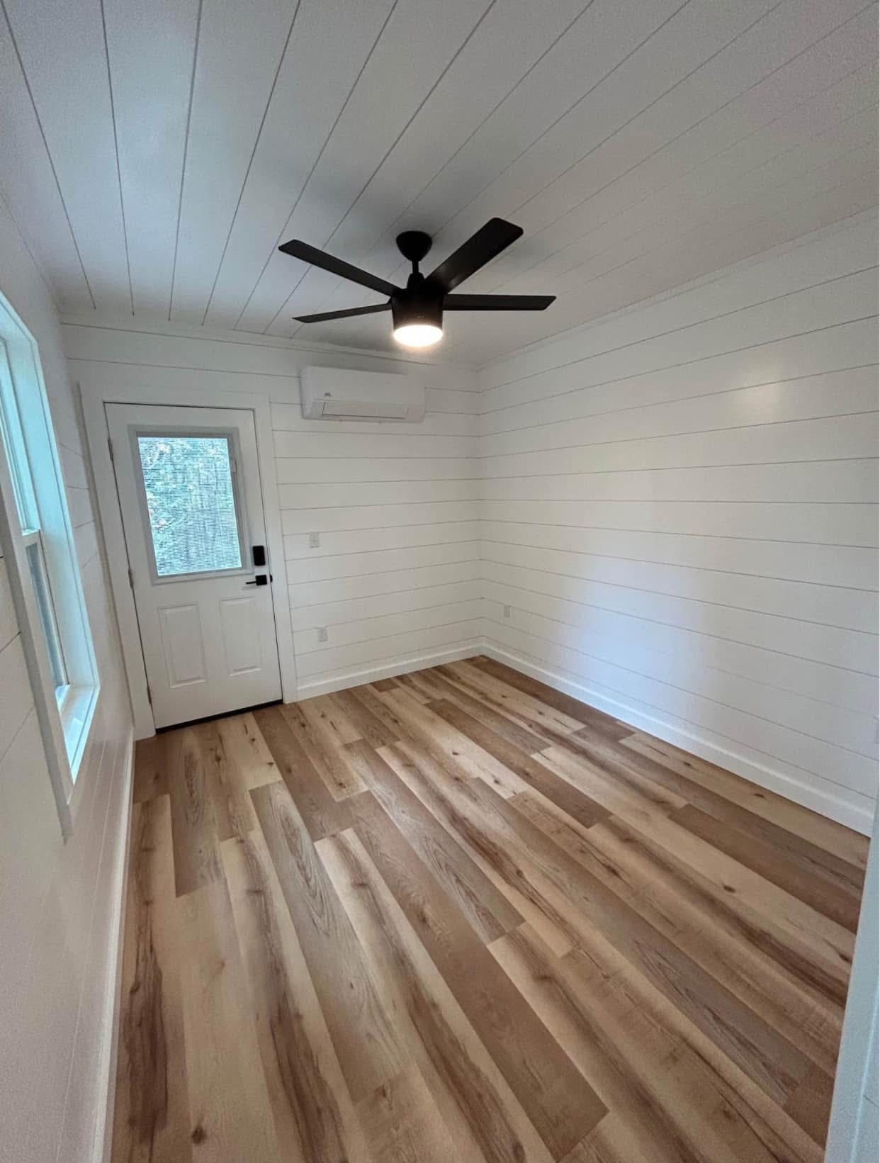 Empty room with white shiplap walls, wood flooring, ceiling fan, and window. Natural light creates a bright, clean, and minimalist atmosphere.
