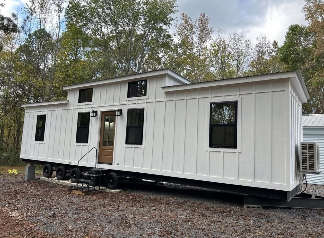 A white mobile tiny house with black-trimmed windows sits on a bed of gravel. It's surrounded by trees, conveying a serene, rustic vibe.