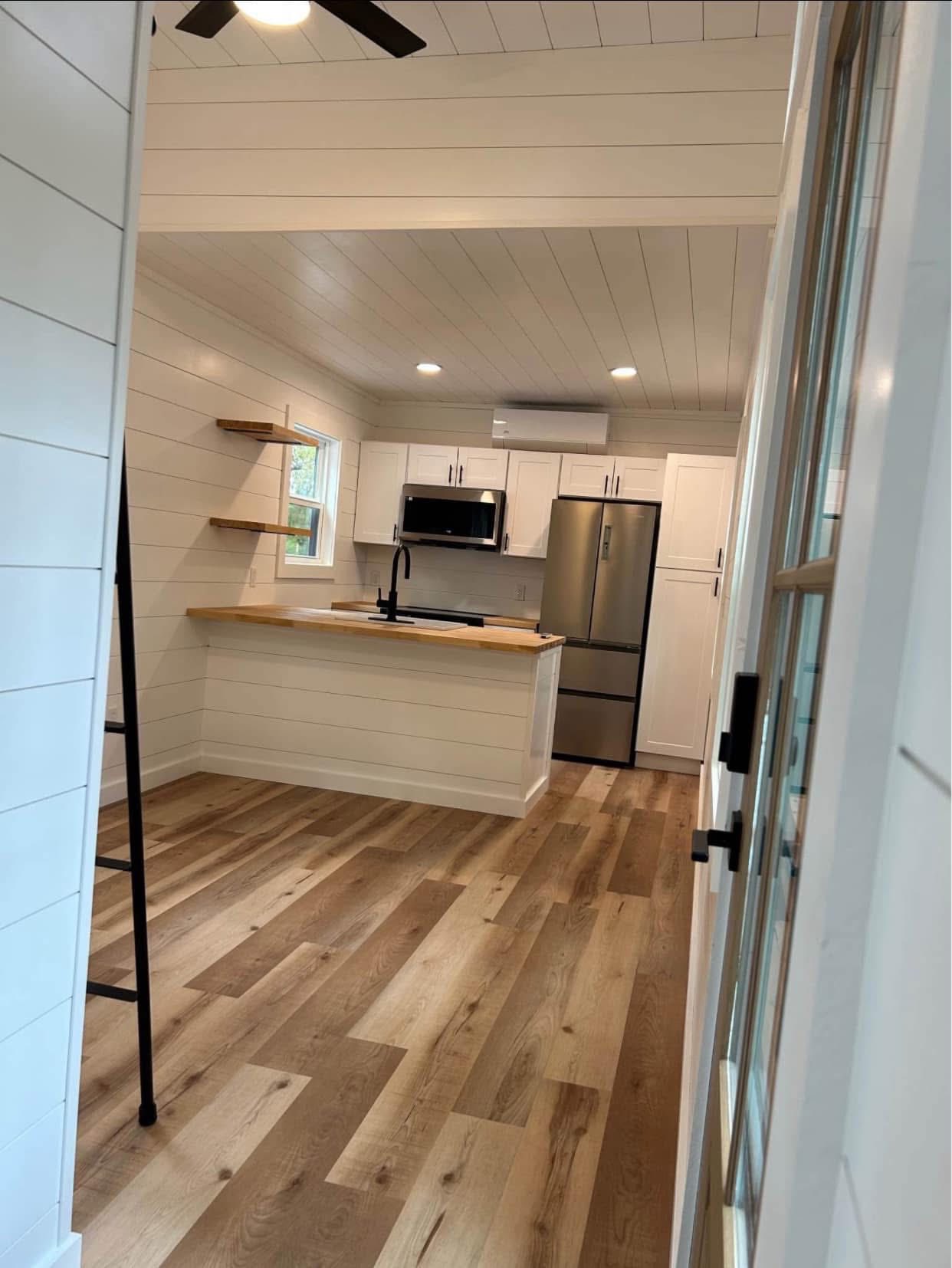 Modern kitchen with white shiplap walls, wood flooring, and a ceiling fan. Stainless steel fridge, microwave, and wood-topped island create a cozy feel.