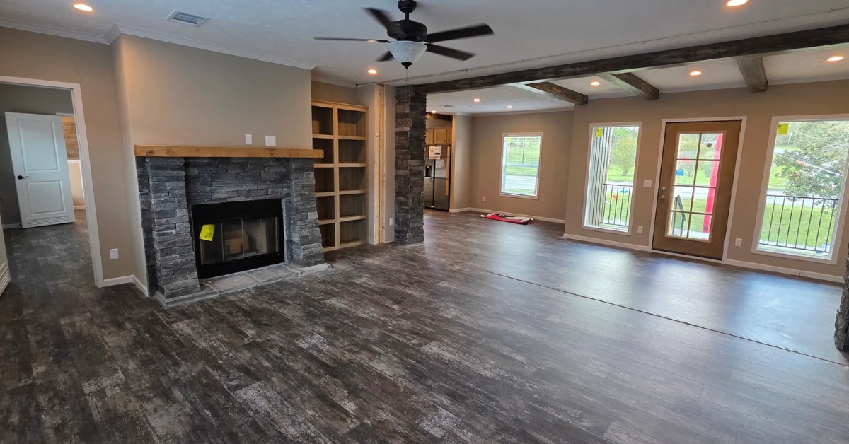 Spacious living room with gray stone fireplace and wooden mantel. Hardwood floors, large windows, ceiling fan, and exposed wooden beams. Bright, inviting space.