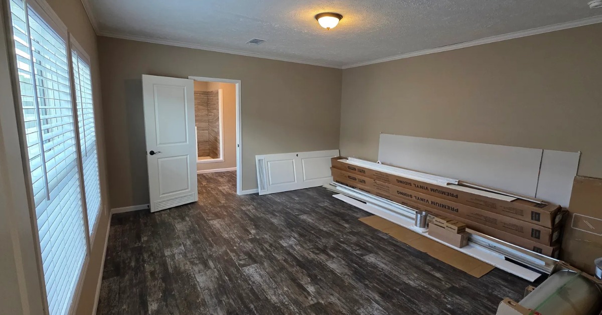 Empty room with wood-look flooring, beige walls, and a single overhead light. White door leads to a bathroom. Stacked cardboard boxes and baseboards suggest renovation.