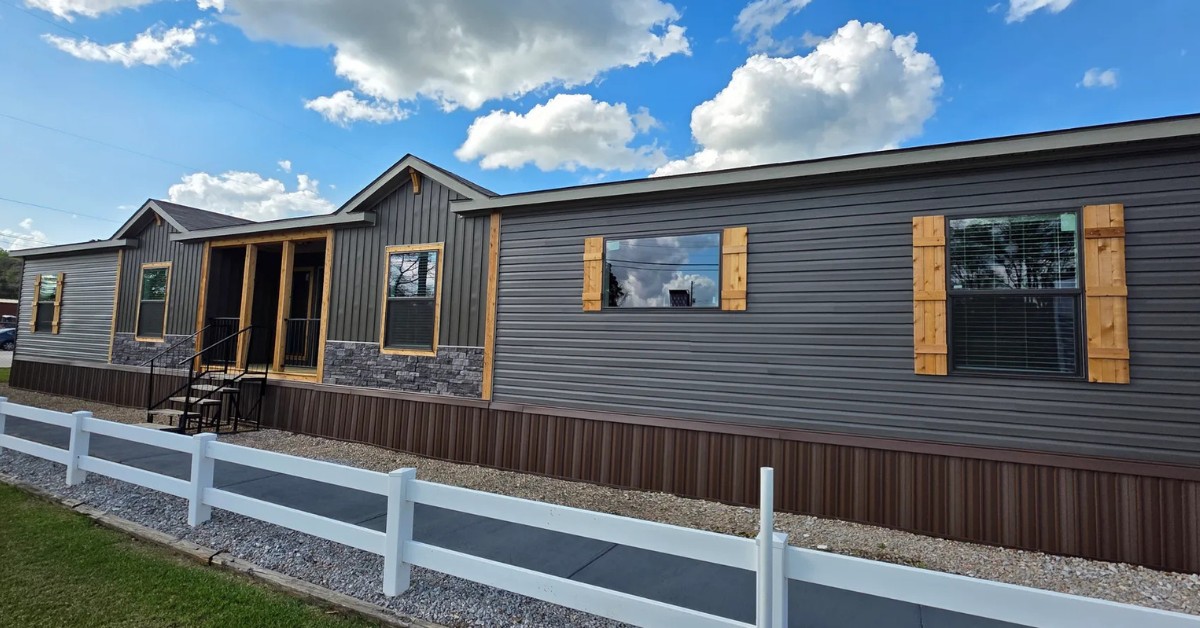 A modern modular home with gray siding and stone accents, featuring wooden shutters. It is set against a backdrop of bright blue sky and fluffy clouds.