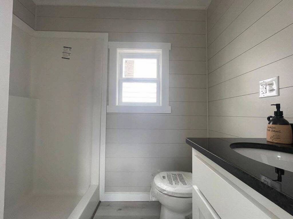 Small, modern bathroom with light gray walls and shiplap design. Features a shower, window, white toilet, and black countertop sink with a soap dispenser. Minimal and clean atmosphere.